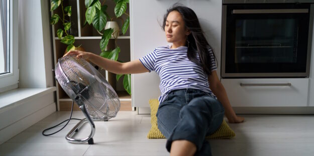 Tired overheated young Asian woman suffering from heat at home woman cooling herself down with a fan