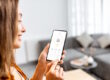 woman in her living room smiling and looking at a smart technology app on her phone